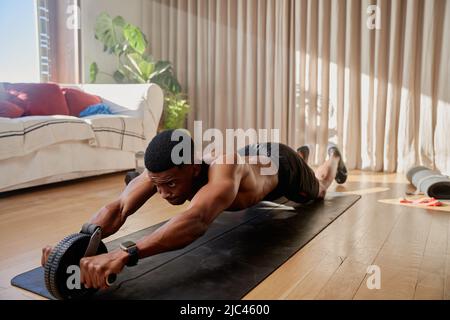 Junger afroamerikanischer schwarzer Mann, der in seinem Wohnzimmer auf seiner Yogamatte trainieren und mit einem Bauchrollerwagen trainieren Stockfoto