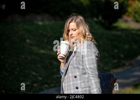 Elegante Blondine mit langen lockigen Haaren in einer leichten Tweed-Jacke, die heißen Kaffee aus einer recycelbaren Tasse trinkt, während sie an einem sonnigen Herbsttag im Park spazieren geht Stockfoto