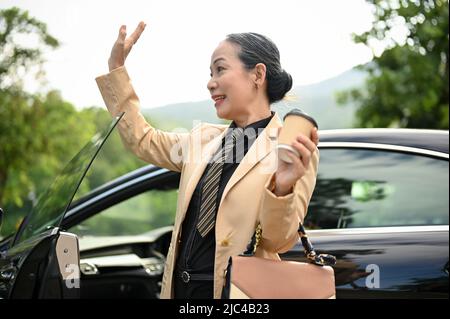 Eine fröhlich gealterte asiatische Geschäftsfrau, die aus ihrem Luxusauto herauskommt und ihre Kollegen mit winkender Hand begrüßt. Stockfoto