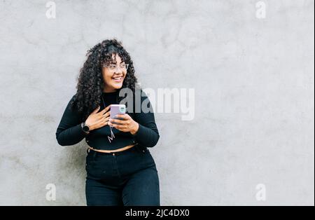 Junge lateinerin glücklich mit ihrem Handy, das an einer Wand gelehnt ist. Eine junge lateinerin, die lachend mit ihrem Handy an einer Wand gelehnt ist, hübsch Stockfoto