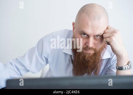 Ein fokussierter Glatze mit einem roten Bart starrt aufmerksam auf die Kamera. Der männliche Manager in einem weißen Hemd ist bei der Arbeit wütend. Stockfoto