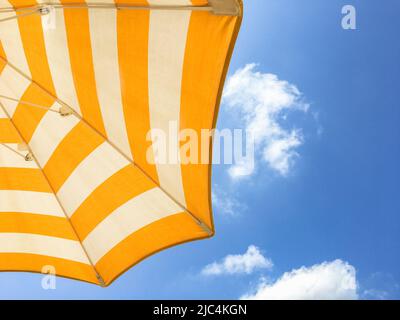 Blick von unten auf den weißen und orangefarbenen Sonnenschirm vor einem blauen Himmel mit weißen Wolken. Urlaubszeit. Stockfoto