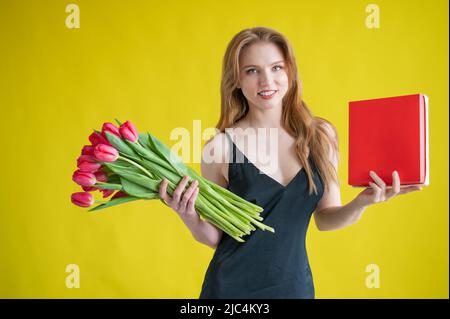 Frau mit einem Strauß roter Tulpen auf gelbem Hintergrund. Glückliches Mädchen in einem schwarzen Kleid hält eine Armvoll Blumen und eine Schachtel Pralinen. Geschenk für Stockfoto
