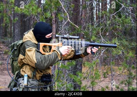 Ein Mann in einer Sturmhaube mit einem Scharfschützengewehr, das in den Wald zielt Stockfoto