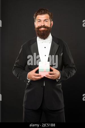 Fröhlicher Mann in Smoking Fliege auf schwarzem Hintergrund mit Geschäft Belohnung Box. Anlass Gruß. Stockfoto