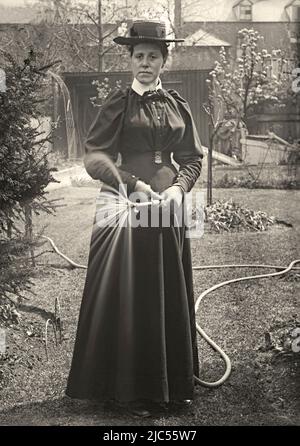 Eine Frau, die einen Schlauch benutzt, um ihren Garten zu bewässern, UK c. 1900. Sie trägt intelligente Kleidung, einschließlich Hut und Handschuhe, für die Aufgabe. Dies ist von einem alten viktorianischen Glasnegativ – einem Vintage 1800s/1900s Foto. Stockfoto