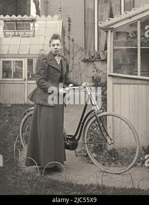 Eine Frau posiert mit dem Fahrrad ihrer Frau in ihrem Garten, Großbritannien c. 1900. Sie trägt geeignete Kleidung zum Reiten. Der Garten hinter dem Haus beherbergt zwei Gewächshäuser. Dies ist von einem alten viktorianischen Glasnegativ – einem Vintage 1800s/1900s Foto. Stockfoto