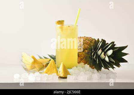 Ananas-Slushie in hohem Glas mit Früchten und zerkleinerten Eis um ihn herum auf weißem Tisch mit isoliertem Hintergrund. Vorderansicht. Horizontale Zusammensetzung. Stockfoto