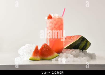 Wassermelone Slushie in hohem Glas mit Früchten und zerkleinerten Eis um sie herum auf weißem Tisch mit isoliertem Hintergrund. Vorderansicht. Horizontale Zusammensetzung. Stockfoto