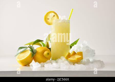 Zitronenschlacken in hohem Glas mit Früchten und zerdrücktem Eis um ihn herum auf einem weißen Tisch mit isoliertem Hintergrund. Vorderansicht. Horizontale Zusammensetzung. Stockfoto