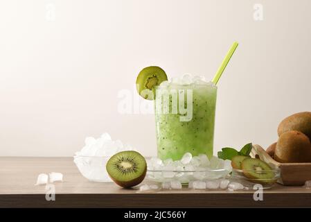 Kiwi Slush trinken im Glas mit Früchten und zerkleinerten Eis um ihn herum auf Holztisch mit isoliertem Hintergrund. Vorderansicht. Horizontale Zusammensetzung. Stockfoto