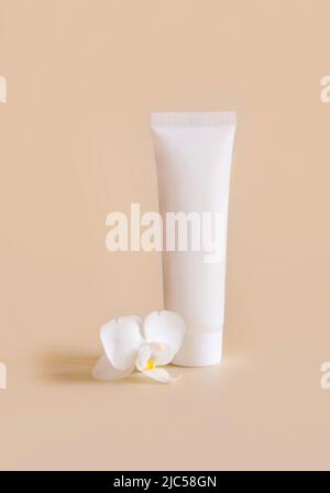 Weiße Plastikcremeröhre in der Nähe der weißen Orchideenblume auf hellgelb, Nahaufnahme, Mockup. Hautpflegeprodukt, Creme oder Lotion. Exotische Naturkosmetik Stockfoto