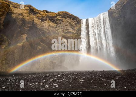 Der mächtige Skógafoss Wasserfall unter einem klaren blauen Himmel, mit einem schönen doppelten Regenbogen, der durch seinen ständigen Sprühnebel verursacht wird, in der Nähe der Route 1 / Ring Road, Souther Stockfoto