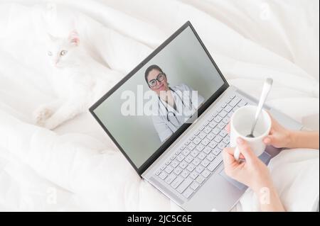 Eine gesichtslose Frau mit Erkältung liegt mit einer Katze im Bett und schaut sich ein Video von einer Arztempfehlung an. Online-Arztbefragung auf einer Webcam ohne Stockfoto