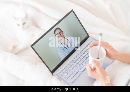 Eine gesichtslose Frau mit Erkältung liegt mit einer Katze im Bett und schaut sich ein Video von einer Arztempfehlung an. Online-Arztbefragung auf einer Webcam ohne Stockfoto