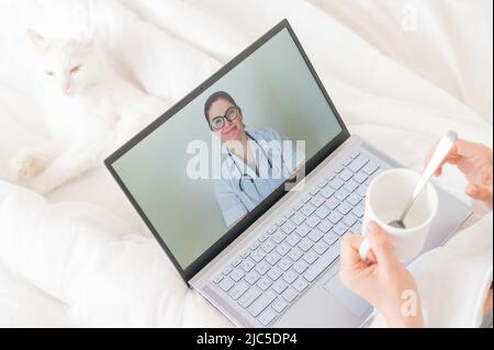Eine gesichtslose Frau mit Erkältung liegt mit einer Katze im Bett und schaut sich ein Video von einer Arztempfehlung an. Online-Arztbefragung auf einer Webcam ohne Stockfoto
