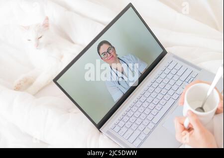 Eine gesichtslose Frau mit Erkältung liegt mit einer Katze im Bett und schaut sich ein Video von einer Arztempfehlung an. Online-Arztbefragung auf einer Webcam ohne Stockfoto
