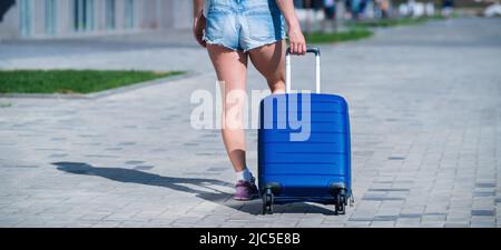 Eine Frau geht und rollt einen blauen Koffer auf Rädern auf dem Bürgersteig. Gepäck ist auf der Straße der Stadt und bereit für die Reise. Rückansicht eines Mädchens in Stockfoto