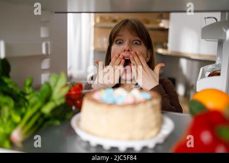 Eine junge Frau blickt überrascht auf einen appetitlichen Kuchen im Kühlschrank Stockfoto