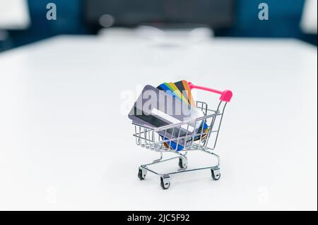 Auf einem weißen Tisch steht ein Mini-Einkaufswagen mit Kreditkarten. Keine Personen. Nahaufnahme eines Wagens mit mehreren verschiedenen Bankkarten. Stockfoto