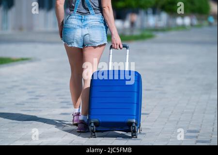 Eine Frau geht und rollt einen blauen Koffer auf Rädern auf dem Bürgersteig. Gepäck ist auf der Straße der Stadt und bereit für die Reise. Rückansicht eines Mädchens in Stockfoto