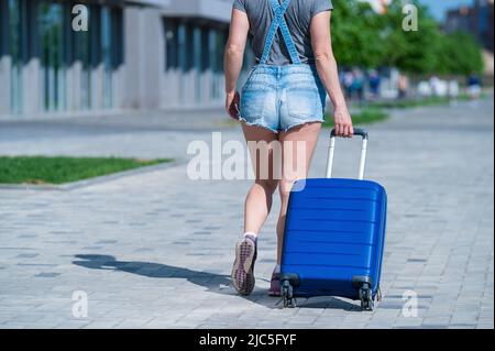 Eine Frau geht und rollt einen blauen Koffer auf Rädern auf dem Bürgersteig. Gepäck ist auf der Straße der Stadt und bereit für die Reise. Rückansicht eines Mädchens in Stockfoto