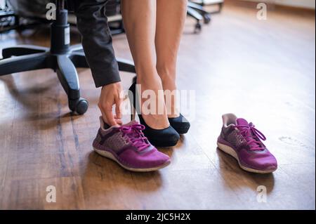 Eine Geschäftsfrau tauscht am Arbeitsplatz High Heels gegen bequeme Schuhe aus. Eine Nahaufnahme weiblicher Hände zieht ihre schwarzen Schuhe aus und zieht farbige an Stockfoto