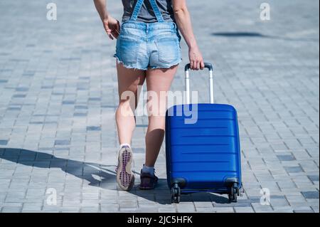 Eine Frau geht und rollt einen blauen Koffer auf Rädern auf dem Bürgersteig. Gepäck ist auf der Straße der Stadt und bereit für die Reise. Rückansicht eines Mädchens in Stockfoto