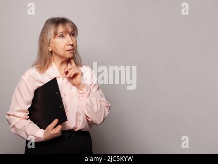 Ältere Geschäftsfrau denkt. Nachdenkliche alte Lehrerin, die mit Brillen auf grauem Banner-Hintergrund wegschaut Stockfoto