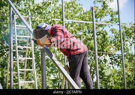 Man Schweißmast mit Schweißmaschine für die Installation von Solarmodulen. Schweißer arbeitet an sonnigen Tag auf dem Feld in Schutzschweißhelm. Metallrohre und Bäume im Hintergrund. Grüne Energie Stockfoto