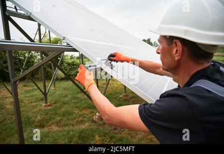 Mann Ingenieur Installation von Solarmodulen. Männlicher Arbeiter in Schutzhelm und Arbeitshandschuhen, die die mittlere Klemme während der Montage des Solar-Photovoltaik-Panelsystems überprüfen. Stockfoto