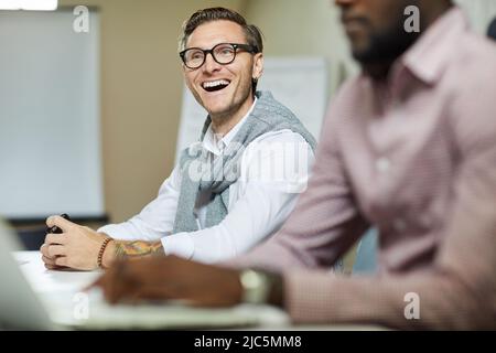 Ekstatisch schöner junger Mann mit Stoppeln, der am Tisch sitzt und beim Treffen im Büro laut lacht Stockfoto