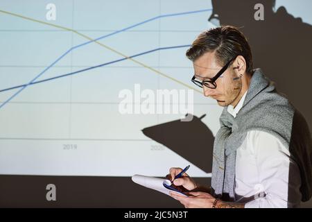 Ernst nachdenklicher junger Lautsprecher mit Stoppeln, der gegen die Projektionsleinwand steht, mit Graphenbild und Aufzeichnungsinformationen in der Zwischenablage Stockfoto