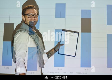 Fröhlicher, aufgeregt junger Mann in einer Brille, die Clipboard hält und Graph auf der Projektionsleinwand kommentiert Stockfoto