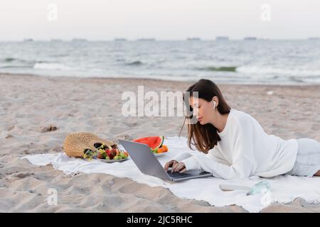 Frau im Kopfhörer mit Laptop in der Nähe des Smartphones und Obst auf der Decke am Strand Stockfoto