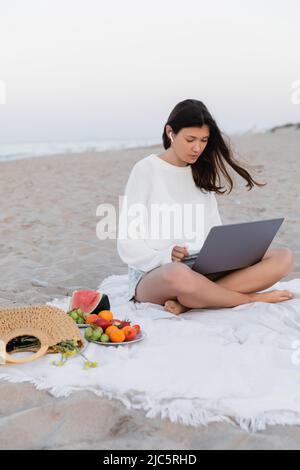 Brünette Frau im Kopfhörer mit Laptop in der Nähe von Früchten auf der Decke Stockfoto