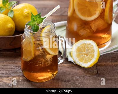 Glas Eistee mit Zitronenscheiben, Minze und Eis auf einem rustikalen Holztisch. Pitcher und Zitronen im Hintergrund Stockfoto