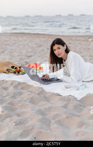 Freiberufler im Kopfhörer mit Laptop in der Nähe von Smartphone und Obst am Strand Stockfoto