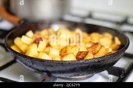 Rösten Sie frische Kartoffeln in einer gusseisernen Pfanne mit Sonnenblumenöl. Ein Blick auf ein Kochfeld mit einer Pfanne gefüllt mit goldenen Bratkartoffeln in einem echten Stockfoto