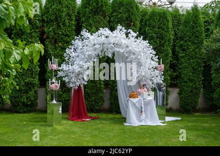 Hochzeitsbogen aus weißen Blumen und anderen Elementen des Hochzeitsdesigns auf dem grünen Rasen. Stockfoto