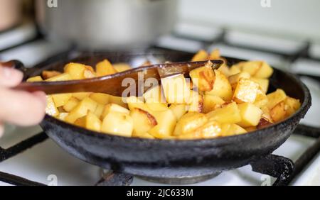 Rösten Sie frische Kartoffeln in einer gusseisernen Pfanne mit Sonnenblumenöl. Ein Blick auf ein Kochfeld mit einer Pfanne gefüllt mit goldenen Bratkartoffeln in einem echten Stockfoto