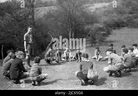 1939, ein Pfadfindermeister mit jungen Pfadfindern, dem 17. Coulsdon, der die Pfadfinderzeremonie des Grand Howl in Devils Den Woods, Dorking, Surrey, England, Großbritannien, durchmachte. In dieser Ära wurden Junge oder Junge Pfadfinder (8-10-Jährige) als Wolf Cubs bekannt. Das Grand Howl, das von Robert Baden-Powell, dem Gründer der Pfadfinderbewegung, entwickelt wurde, basiert auf dem Dschungelbuch', einem Roman von Rudyard Kipling, in dem ein Wolfspackentreffen beschrieben wird, das damit beginnt, dass jeder seinen Anführer umkreist und heult. Beim Scouting wurde dies zum 'Grand Howl', mit der Idee, dass sich die jungen Scouts versammeln und sich konzentrieren. Stockfoto