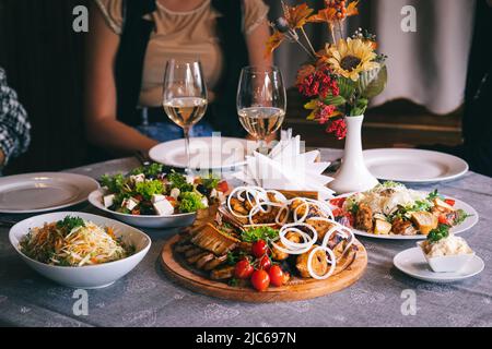 Teller mit Grill, Sauce und Salaten. Abendessen im Restaurant. Im Hintergrund sind Gläser Weißwein zu sehen. Stockfoto