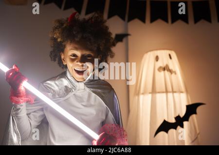 Portrait eines positiv ekstatischen afroamerikanischen Jungen mit offenem Mund, der glühenden Lichtstab hält, Halloween Party Konzept Stockfoto