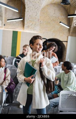 Glückliche Frau mit Copybooks, die in der Nähe verschwommener interrassischer Freunde im Klassenzimmer wegschauen Stockfoto