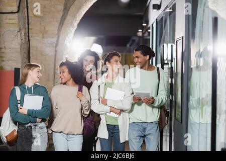 Fröhliche multikulturelle Studenten mit Rucksäcken und Gadgets, die durch den Korridor laufen Stockfoto