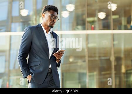Seriös nachdenklicher, gut gekleideter afro-amerikanischer Geschäftsmann, der gegen Bürogebäude steht und ein modernes Telefon nutzt Stockfoto