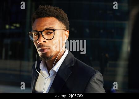 Porträt eines seriös hübschen jungen bärtigen schwarzen Geschäftsmannes in einer stilvollen Brille, der in Kopfhörern Radio-Nachrichten hört Stockfoto