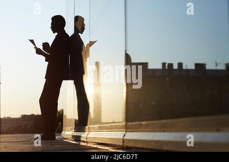 Silhouette eines Mannes im Anzug, der neben einem glasigen Gebäude steht und Kaffee trinkt, während er im Freien Papiere analysiert, Reflexionseffekt Stockfoto