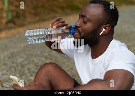 Junger erwachsener Mann, der sich nach einem Lauf mit einer Flasche Wasser und einem Smartphone auf einem Park ausruhte. Workout im Freien Stockfoto